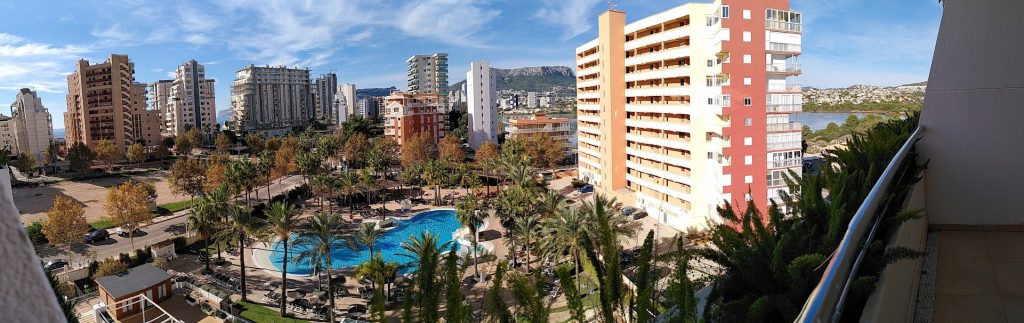 A panarama shot from the balcony of the pool showing a blue pool surrounded by palm trees, a number of high rise building and in the distance, mountains.